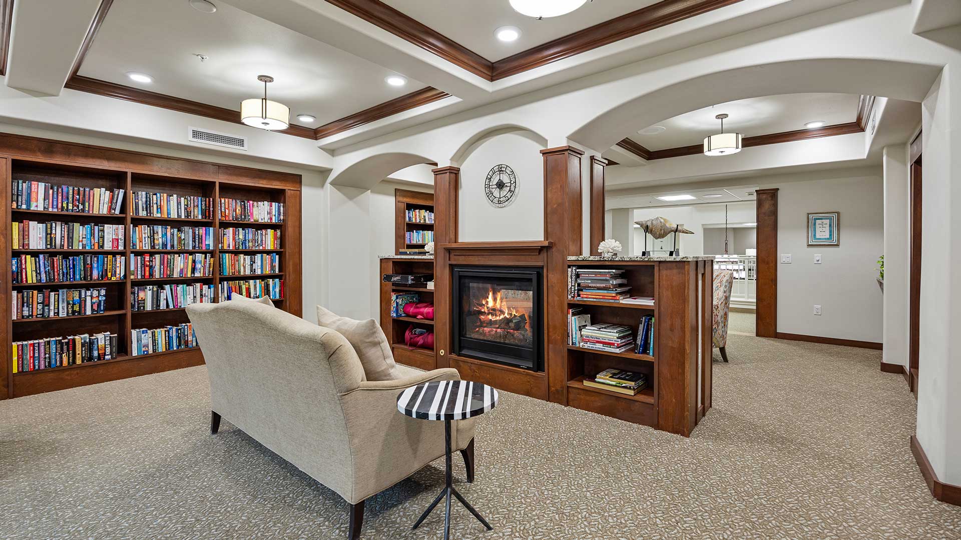 A cozy room with a beige sofa facing a fireplace, surrounded by wooden shelves filled with books. The ceiling has recessed lighting and elegant moldings. A small round table is beside the sofa, and a clock is mounted above the fireplace.