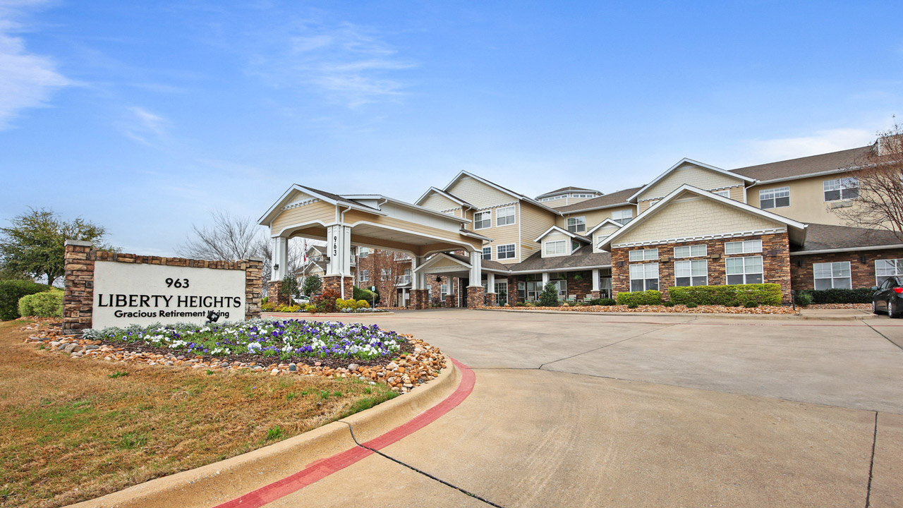 A large retirement home with beige and white exterior, tall columns at the entrance, manicured landscaping, and a sign reading "Liberty Heights Gracious Retirement 963" near the driveway. Clear blue sky above.