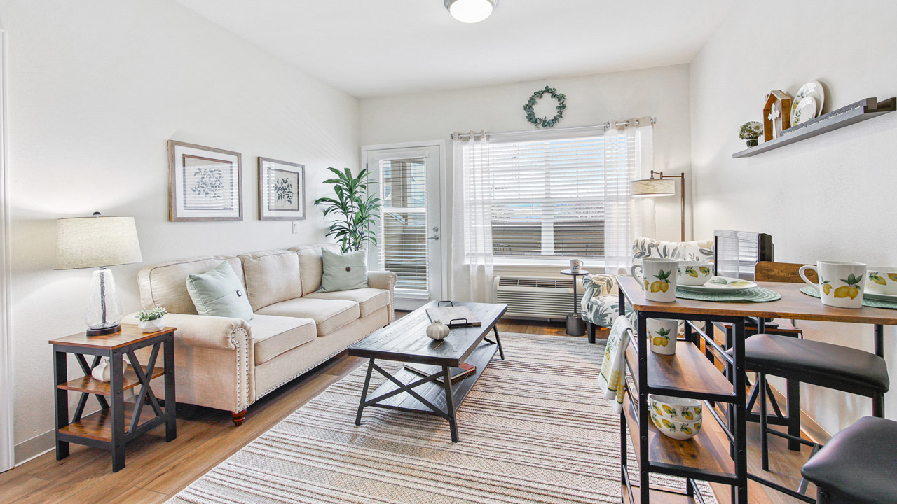 Bright, cozy living room with a beige sofa, coffee table, side tables with lamps, wall art, a rug, a small dining table with chairs, and a window letting in natural light. Plants and decorative items add warmth to the space.