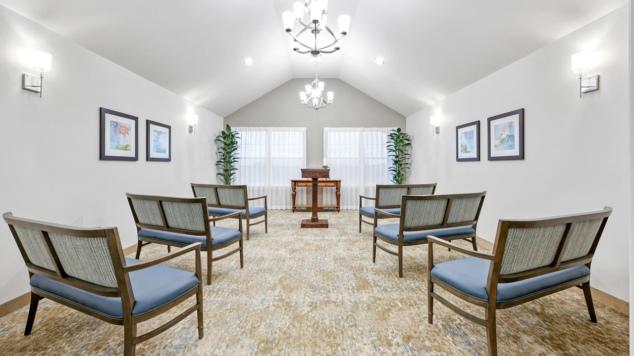 A small, bright chapel room with six chairs arranged in two rows facing a wooden podium, framed artwork on white walls, two tall plants, carpeted floor, and large windows with sheer curtains.