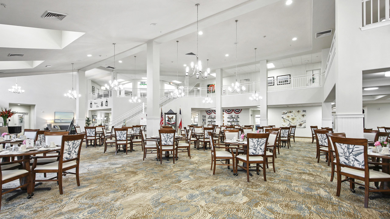 Spacious, elegant dining hall with wooden tables and chairs, chandeliers, white walls, and a balcony. Table settings and decor create a welcoming atmosphere, with natural light from high ceilings and large windows.