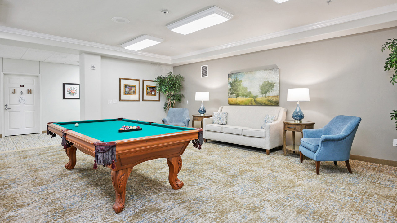 A bright, spacious room with a pool table, two blue armchairs, a beige sofa, lamps, framed artwork, and a potted plant on a neutral-patterned carpet. The atmosphere is cozy and inviting.