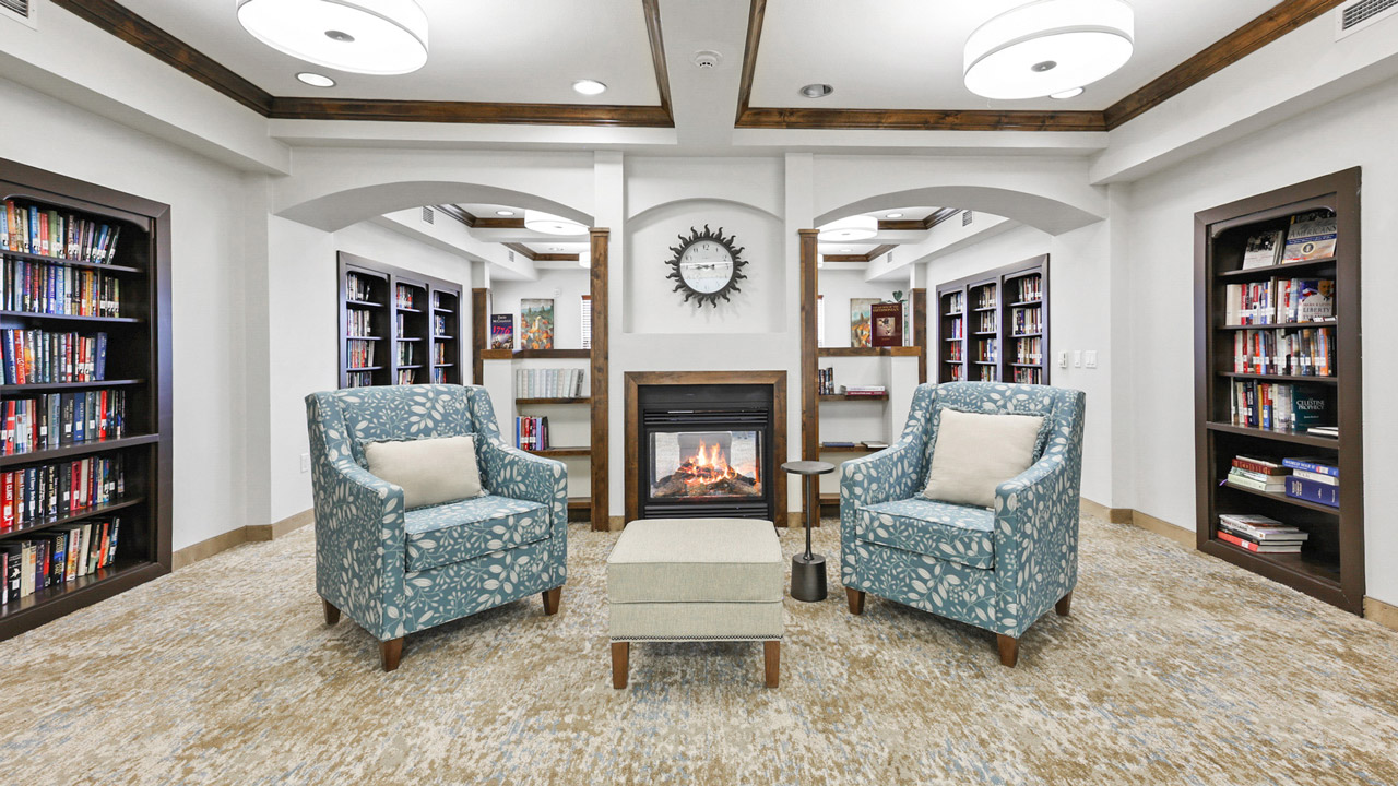 Two blue patterned armchairs and a small ottoman face a fireplace with a clock above it, surrounded by bookshelves in a cozy, well-lit library room with neutral décor.