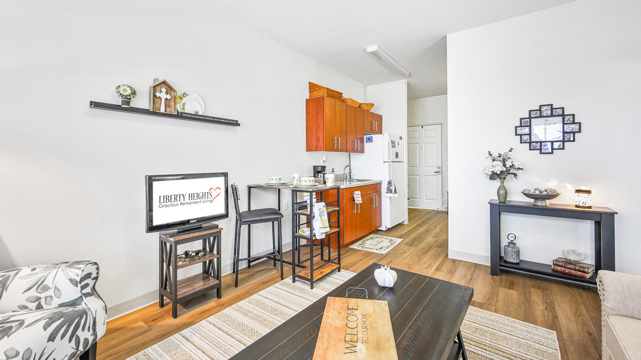 A cozy apartment living area with wood flooring, a small kitchen, a TV on a stand, two barstools at a counter, a sofa, a coffee table, and decorative items on shelves and a side table against white walls.