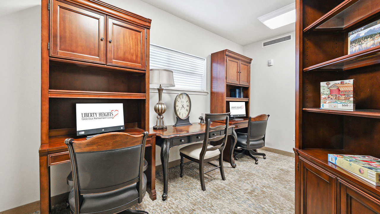 A home office with two wooden desks and chairs, each with a computer monitor. There is a clock and lamp on the center desk. Bookshelves line the walls, holding board games and books. A window lets in natural light.