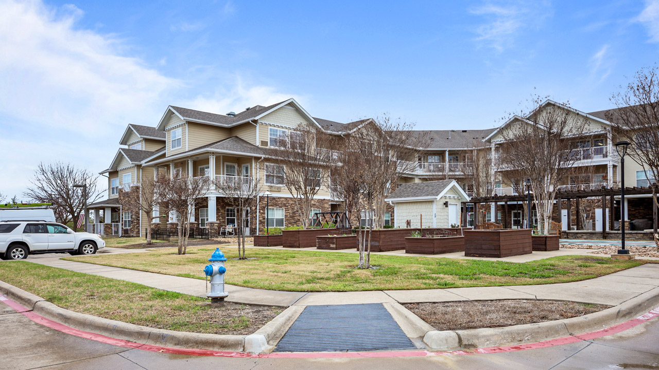 A three-story residential building with balconies, raised garden beds, leafless trees, a white SUV, a blue fire hydrant, and a sidewalk under a partly cloudy sky.