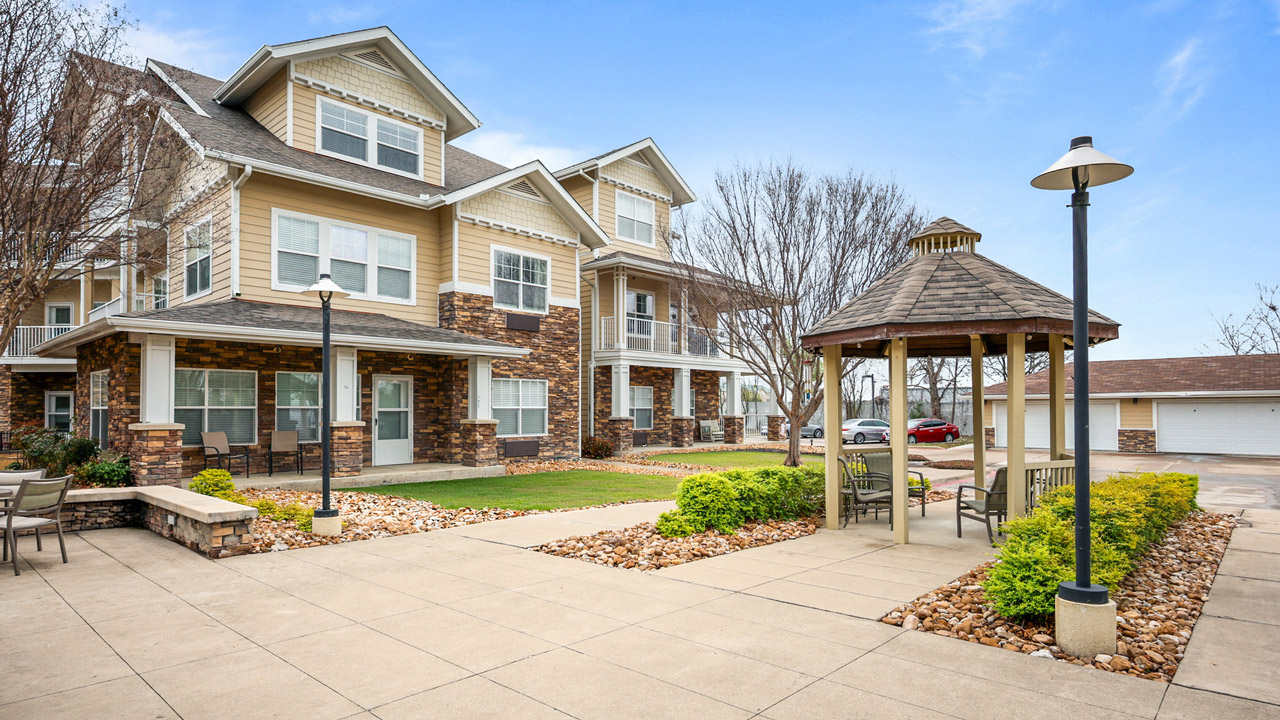 A modern apartment building with stone and beige siding, landscaped gardens, a gazebo with benches, and paved walkways under a clear blue sky.