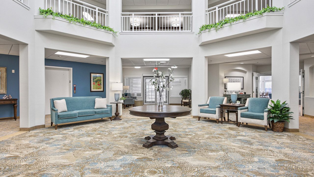 Spacious, bright lobby with a round wooden table and flower vase at the center, blue armchairs and sofas, potted plants, and white railings on the upper level. Light decor and patterned carpet create a welcoming atmosphere.