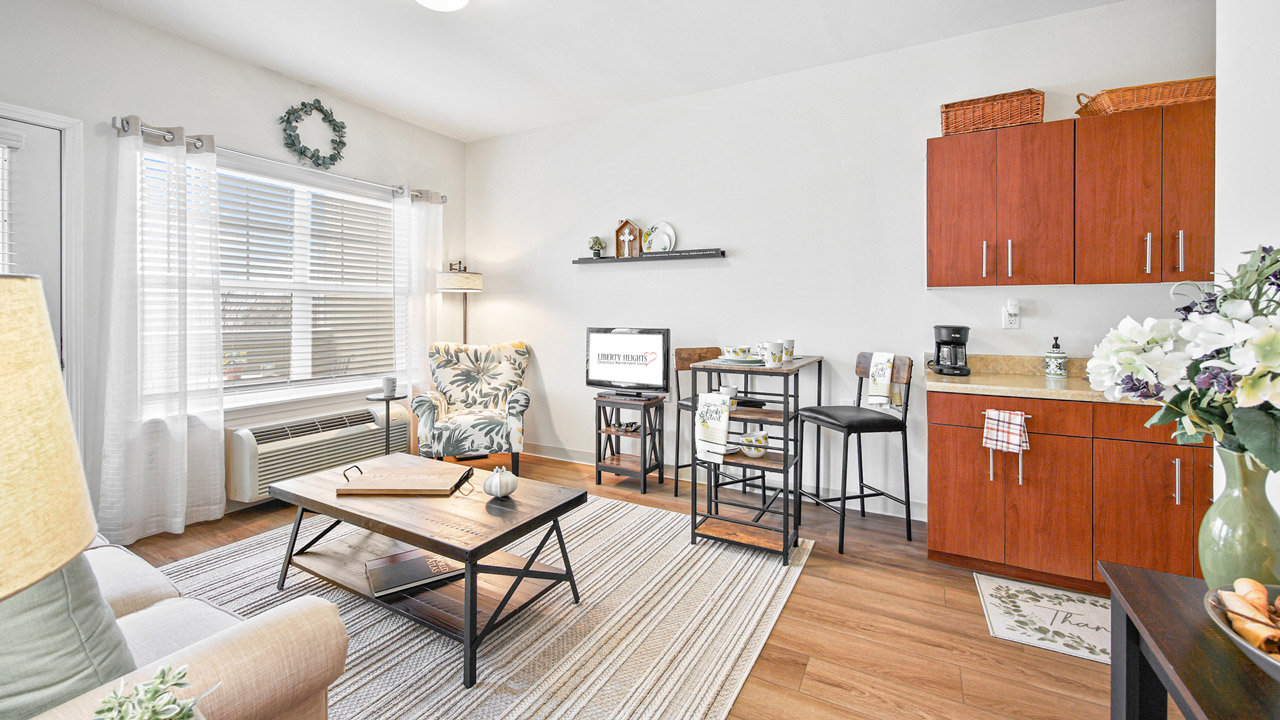A bright, cozy living space with a sofa, coffee table, TV, and a small dining area. Wood floors and cabinetry, large windows with blinds, and decorative accents create a welcoming, modern atmosphere.