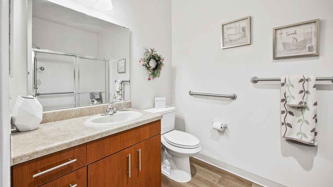 A modern bathroom with a wood vanity, white sink, toilet, wall-mounted towel rack, shower with glass doors, and decorative elements including framed art and a floral wreath. The decor is minimalist and clean.