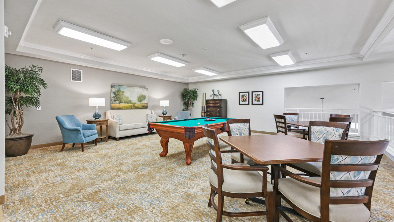 A spacious game room features a pool table, a table with four chairs, a sofa, armchairs, potted plants, wall art, and bright ceiling lights, creating a comfortable and inviting atmosphere.