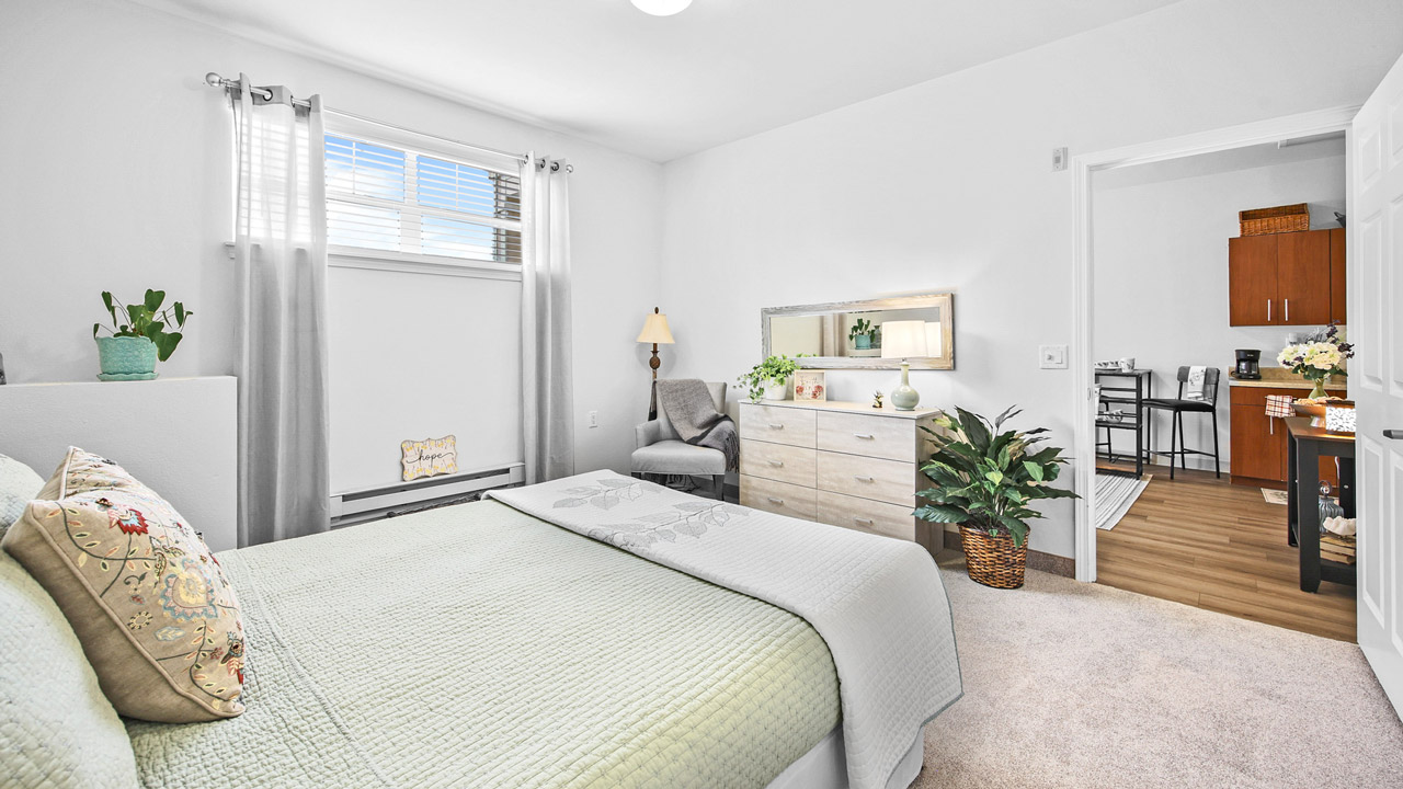 Bright bedroom with a neatly made bed, pale green bedding, dresser with a mirror, plants, and decor. Doorway leads to a kitchen with wood floors and dark cabinets. Sunlight streams through a window with sheer curtains.