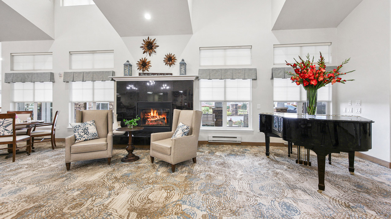 A spacious, elegant living room with a fireplace, two beige armchairs, a small table, a black grand piano with a large flower arrangement, and large windows letting in natural light.