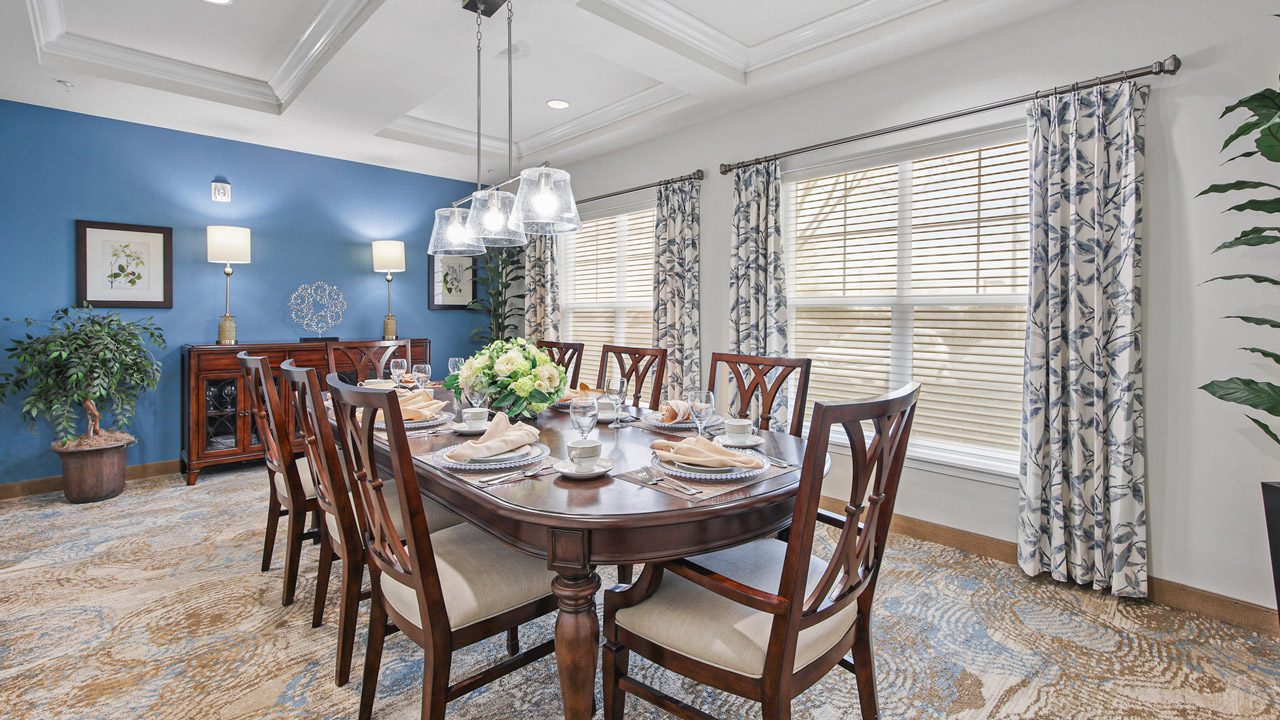 Elegant dining room with a dark wood table set for eight, upholstered chairs, a floral centerpiece, blue accent wall, patterned curtains, large windows, carpeted floor, and decorative artwork and plants.