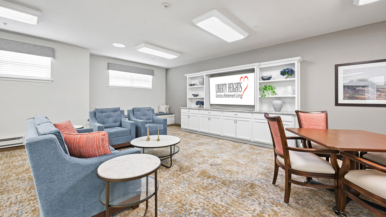 A bright, modern lounge with blue armchairs, a white sofa, a round coffee table, and a wooden dining table with chairs. Built-in shelves and a TV display "LIBERTY HEIGHTS, Cotton Extended Living" on the far wall.