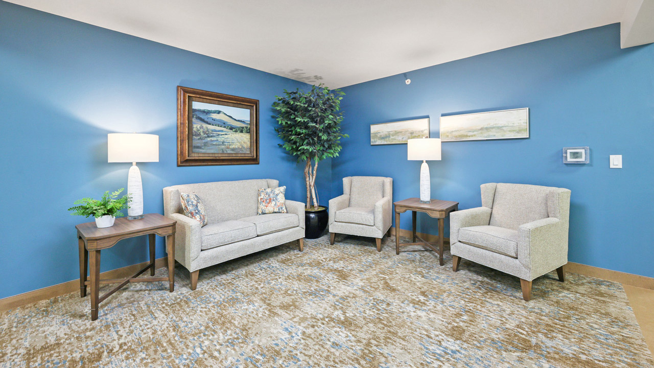 A cozy seating area with two beige armchairs and a loveseat, two side tables with lamps, a potted plant, and landscape artwork on blue walls over a patterned carpet.