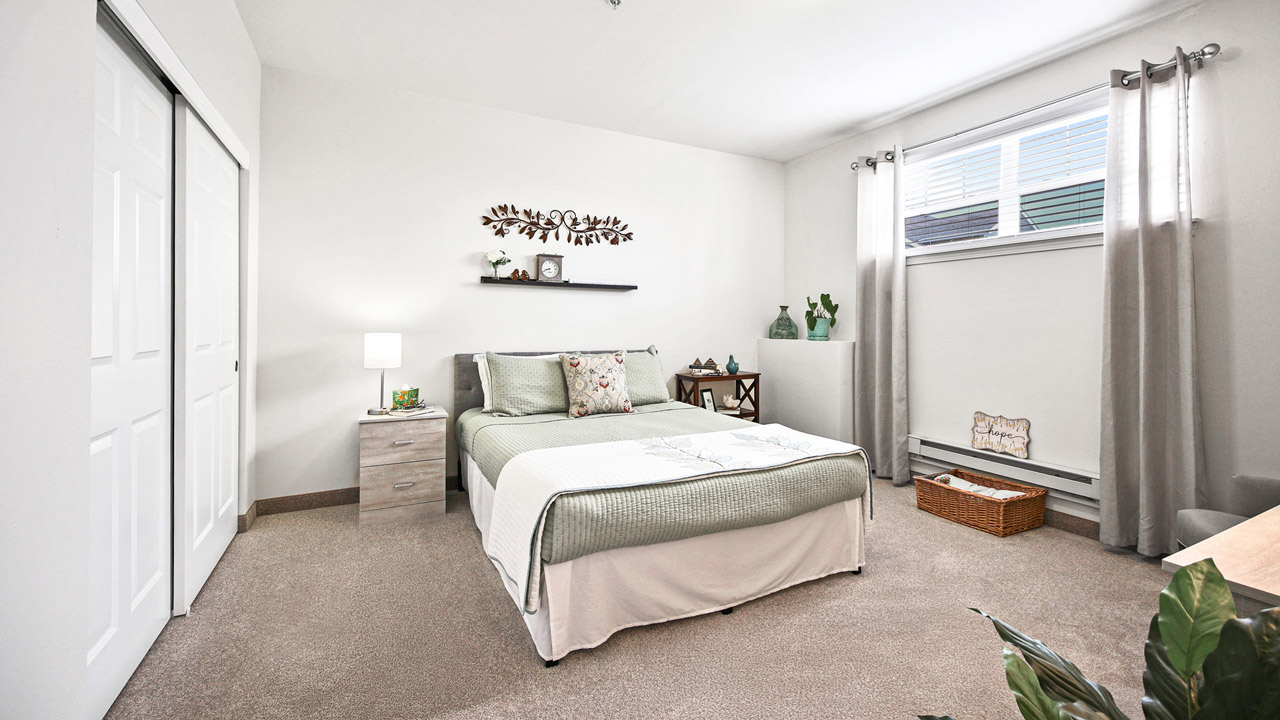 A bright, modern bedroom with a neatly made bed, light-colored bedding, nightstand with a lamp, wall art, plants, large window with sheer curtains, and neutral decor.