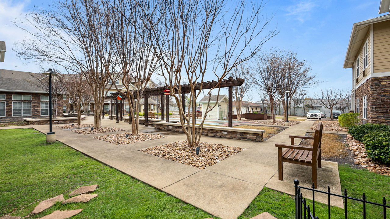 A landscaped courtyard features leafless trees, a pergola with benches, stone pathways, and green grass. Residential buildings and a parked car are visible in the background under a blue sky.