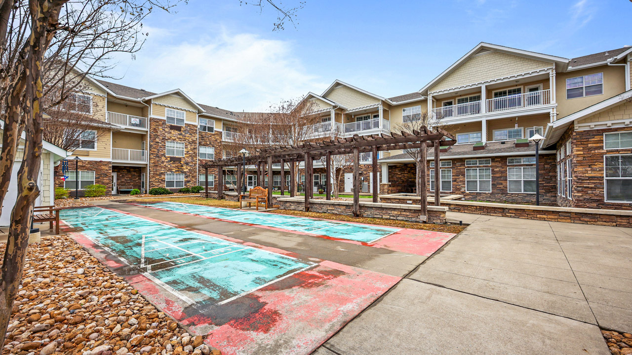 A courtyard with a pergola, benches, and two shuffleboard courts, surrounded by a three-story apartment building with stone and beige siding, under a partly cloudy sky.