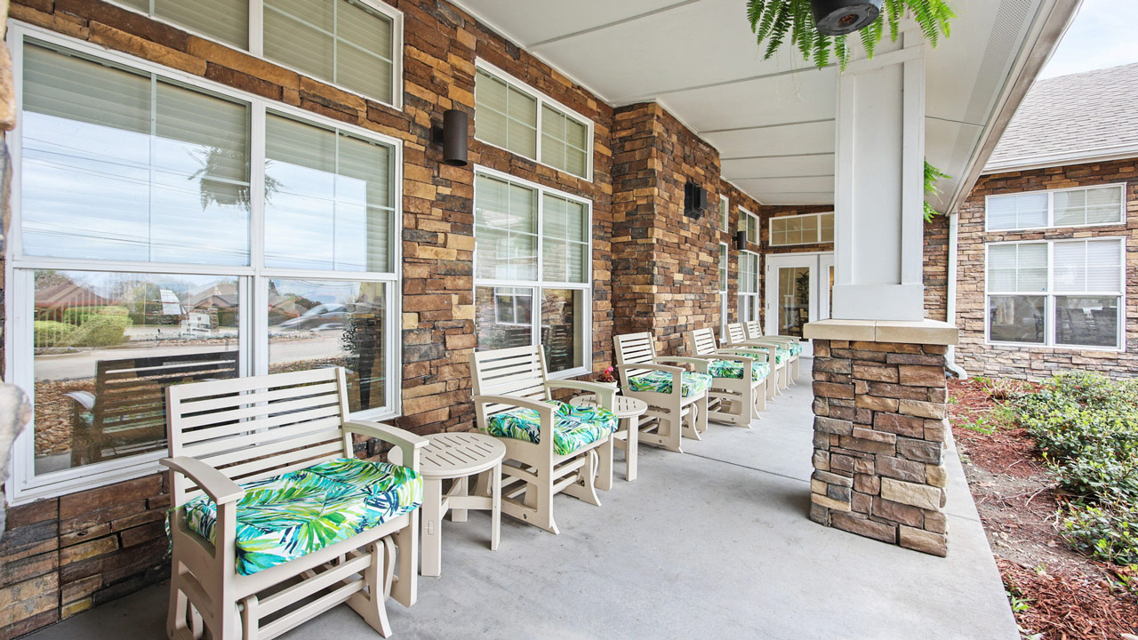 A covered porch with several cream-colored chairs featuring green patterned cushions, matching side tables, and stone walls. Large windows line the building, and ferns hang from the ceiling.