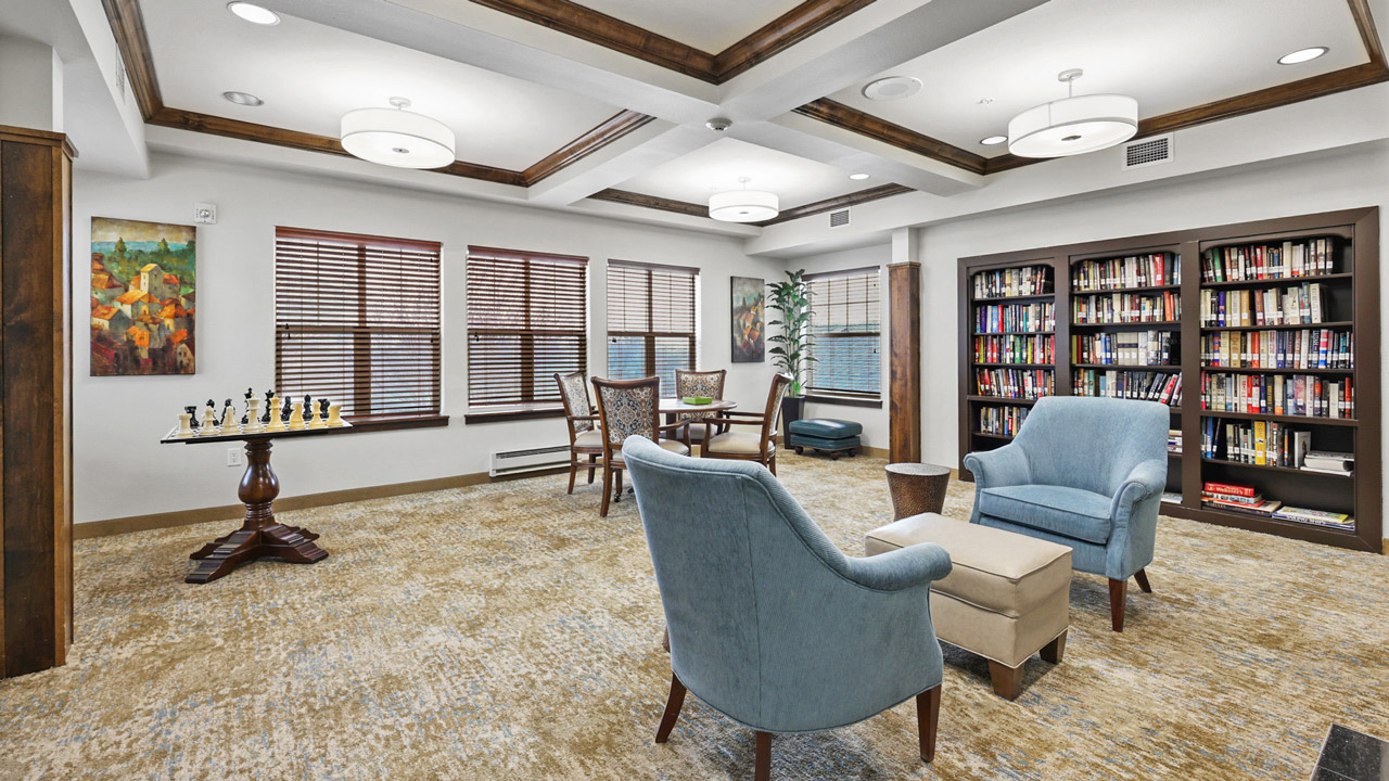 A cozy library room with two blue armchairs, a small ottoman, a round wooden table, bookshelves filled with books, a chess table, artwork on the wall, and large windows with blinds letting in natural light.
