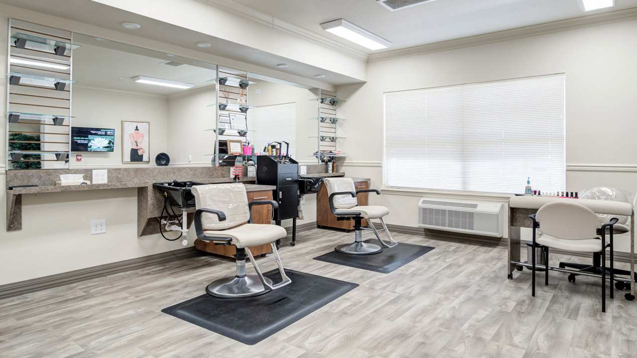 A clean, modern salon with two empty styling chairs facing a large mirror, shelves with hair products, and a manicure table with nail polish. The room has bright lighting, wood-style flooring, and a window with closed blinds.