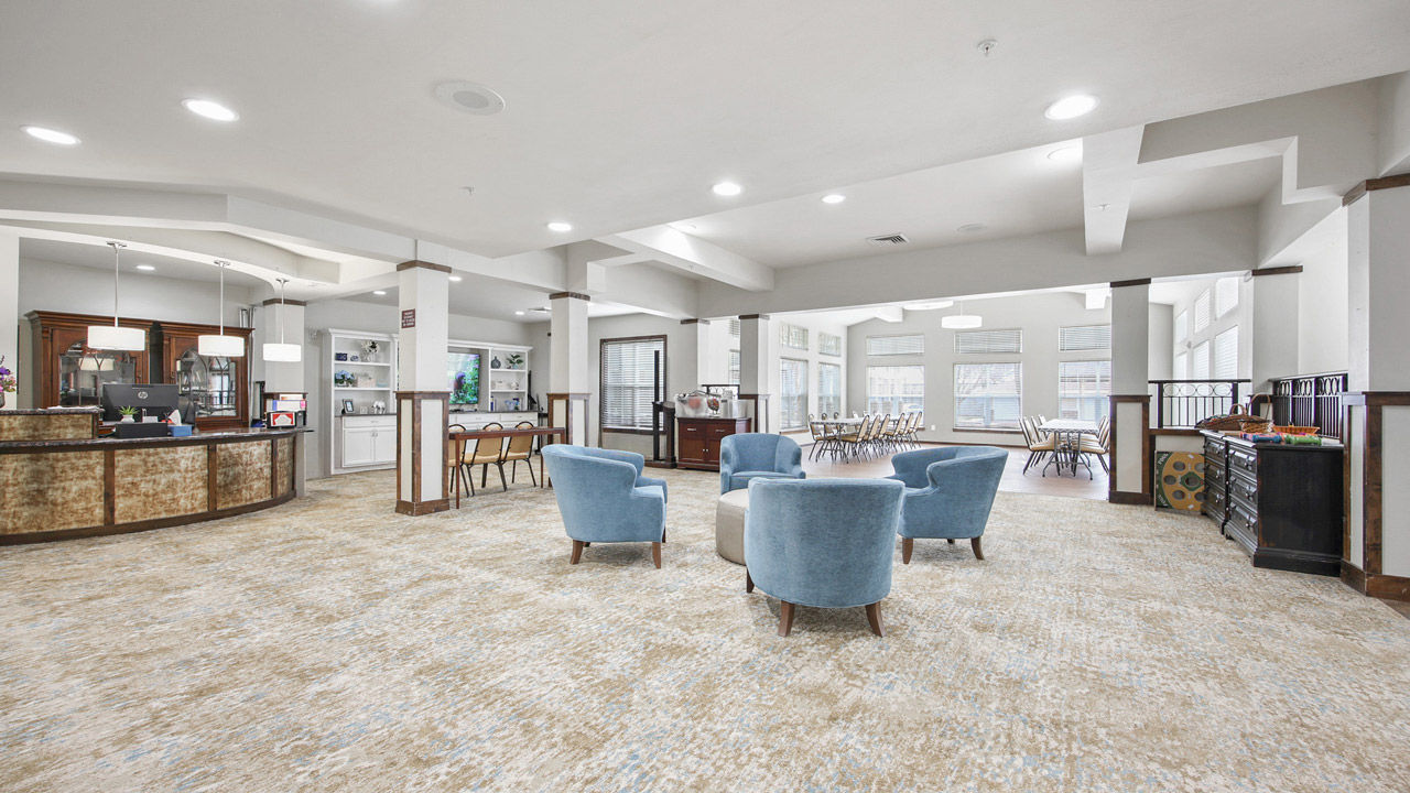 A spacious, modern lounge area with four blue chairs around a small table, a reception desk on the left, and large windows letting in natural light at the back. Dining tables are visible in the background.