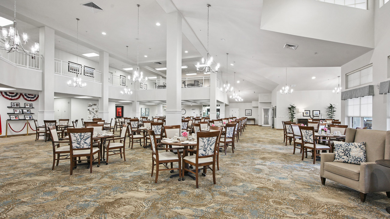 Spacious, elegant dining room with multiple wooden tables and chairs, chandeliers, large windows, neutral décor, a high ceiling, and patriotic bunting on the far wall. Some tables are set with dishes and pink flowers.