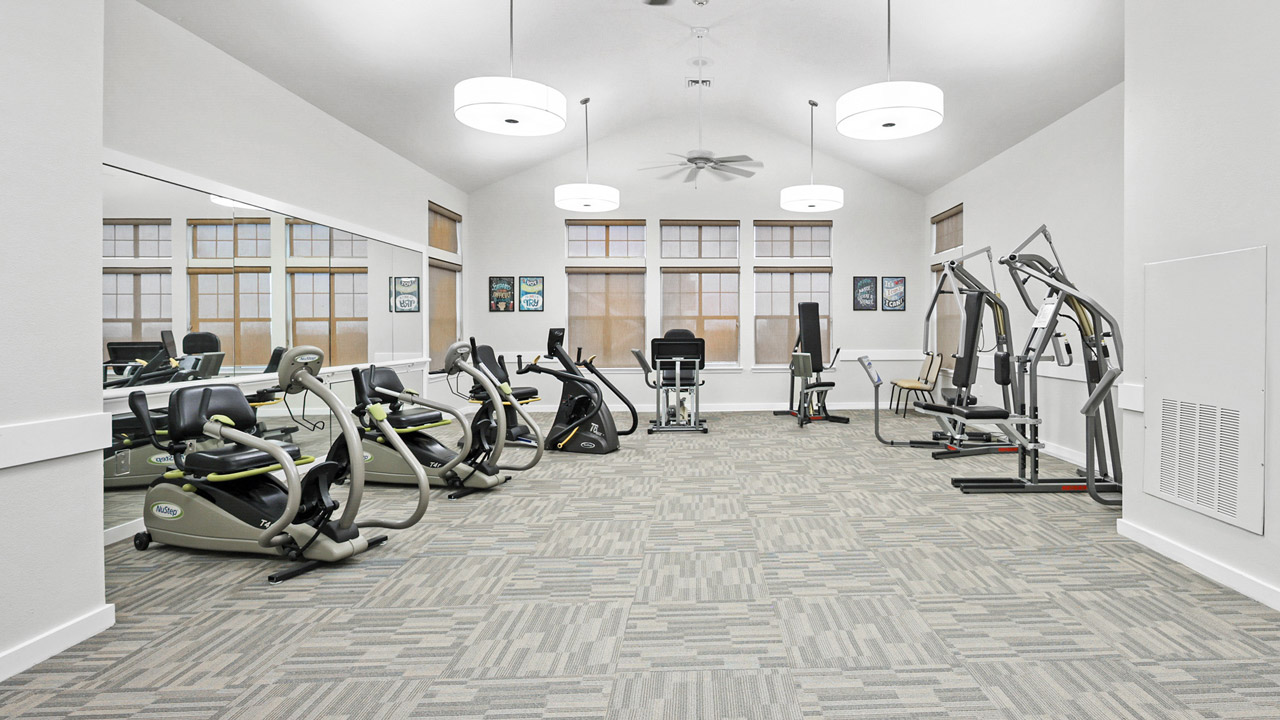 A bright, spacious gym with exercise machines, including recumbent bikes, an elliptical trainer, and strength training equipment, arranged on patterned carpet with large windows and wall mirrors.