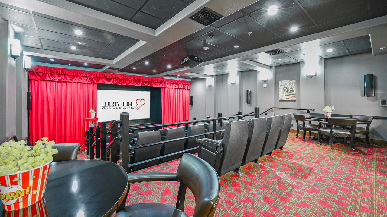 A small movie theater with rows of black seats, red carpet, and a screen displaying "Liberty Heights Gracious Retirement Living." Red curtains frame the screen, and chairs with tables and popcorn are in the foreground.