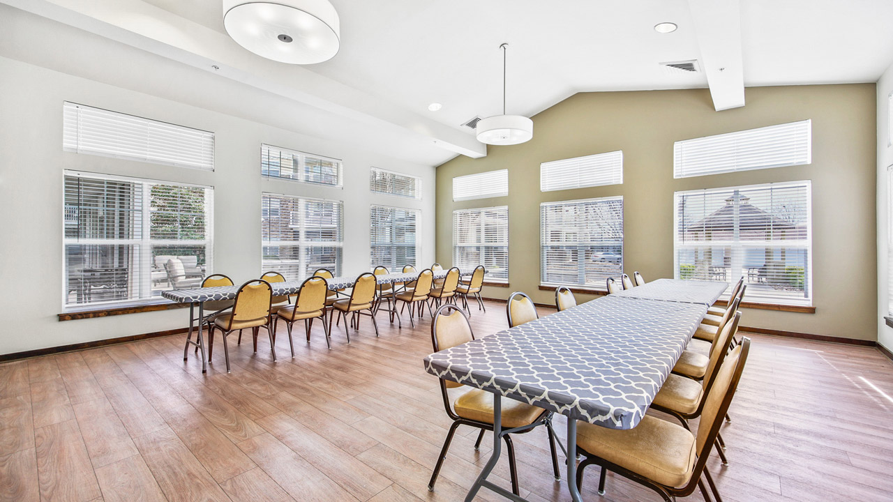 A bright, spacious room with light wood floors, large windows, and long tables covered with patterned gray and white tablecloths. Yellow chairs are arranged around the tables. The room appears set up for a meeting or gathering.