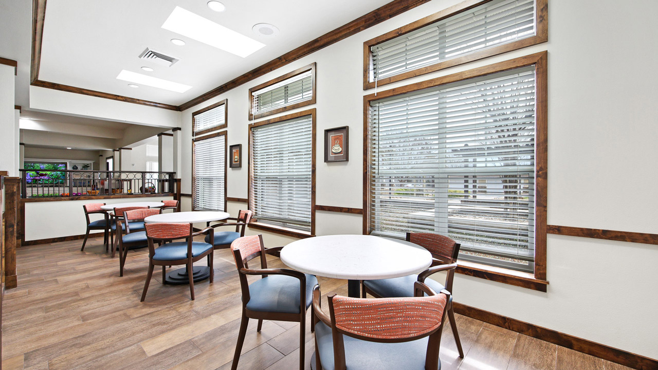 A bright, modern room with several round tables and chairs on a wooden floor, large windows with blinds, and framed artwork on the walls. Natural light fills the space, creating an inviting atmosphere.