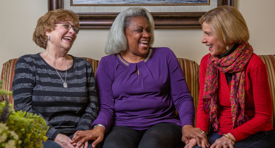 Three older women are sitting on a striped couch, laughing together. The woman on the left wears glasses and a striped sweater, the woman in the middle has silver hair and wears a purple top, and the woman on the right wears a red sweater and a colorful scarf.