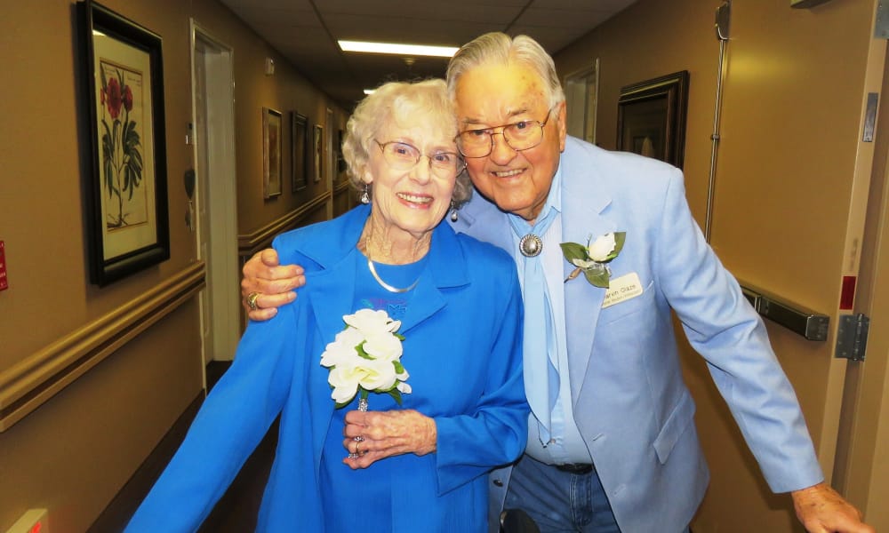 An elderly couple wearing matching blue outfits stands in a hallway, smiling warmly at the camera. The man has his arm around the woman, who is holding a white flower bouquet. The hallway features framed artwork on the walls.