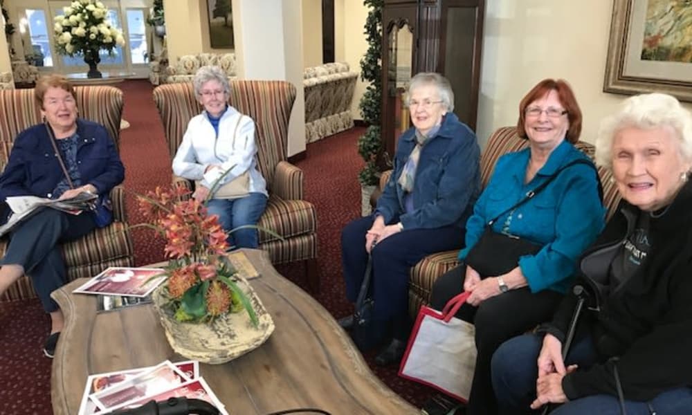 Five elderly women are sitting together in a cozy living room, smiling at the camera. They are seated on armchairs and a couch, surrounded by warm decor and a wooden coffee table with some magazines. The atmosphere appears friendly and inviting.