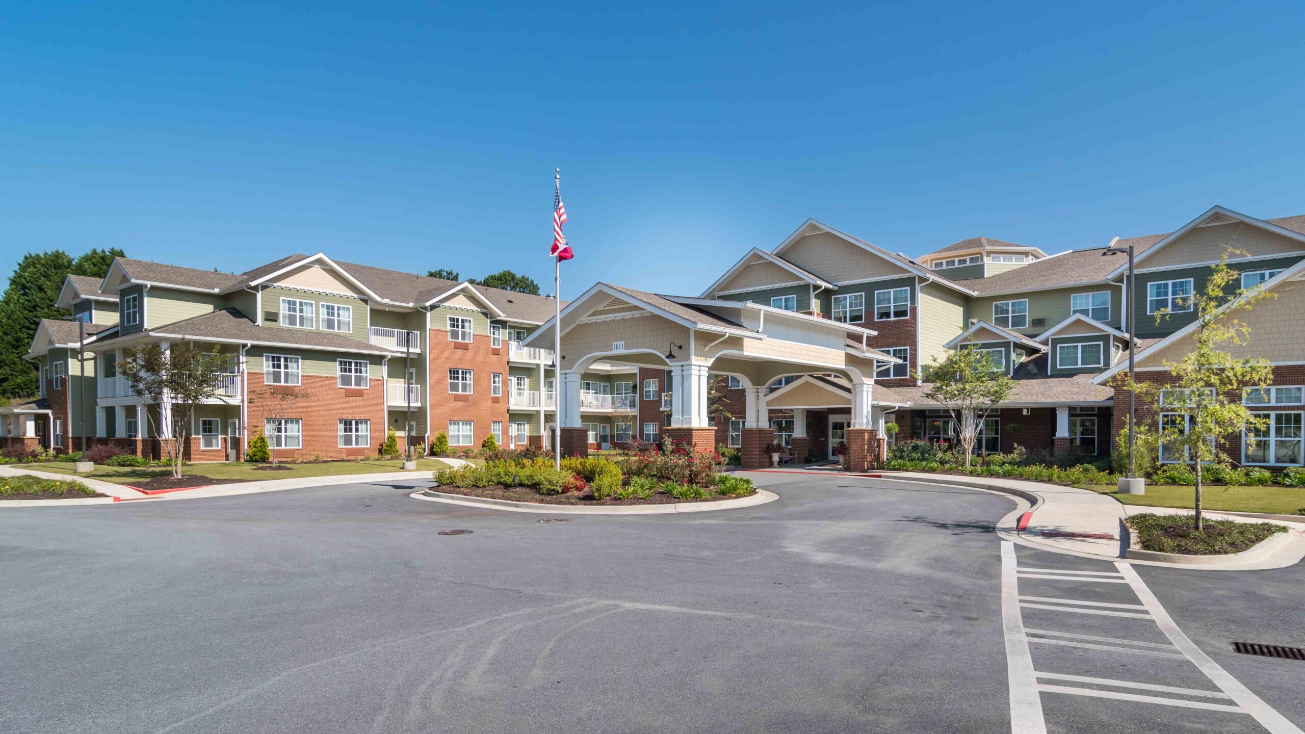A large, modern assisted living facility with multiple stories, landscaped gardens, and a circular driveway; an American flag is raised on a flagpole in front of the main entrance under a clear blue sky.