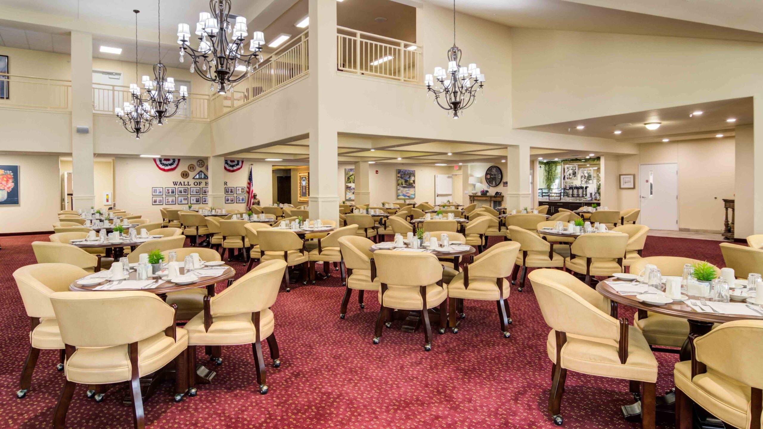 A spacious dining room with round tables set for meals, cream-colored chairs, red carpet, chandeliers, and patriotic decorations on the walls. There is a balcony area above and framed pictures on display.