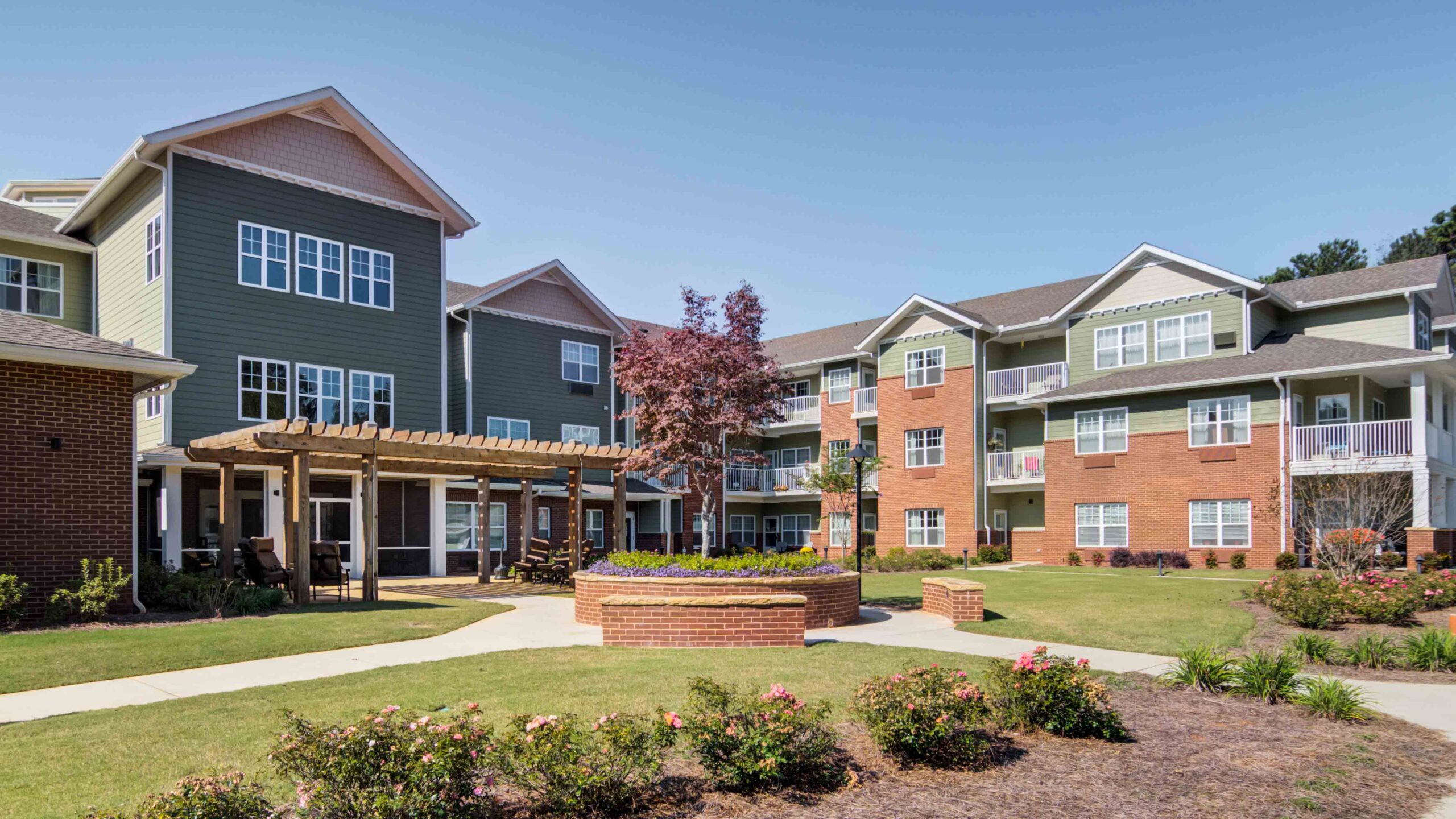 A modern apartment complex with three-story buildings, balconies, and large windows. There’s a central landscaped courtyard with a round flower bed, a tree, a wooden pergola, and walking paths surrounded by green lawns.