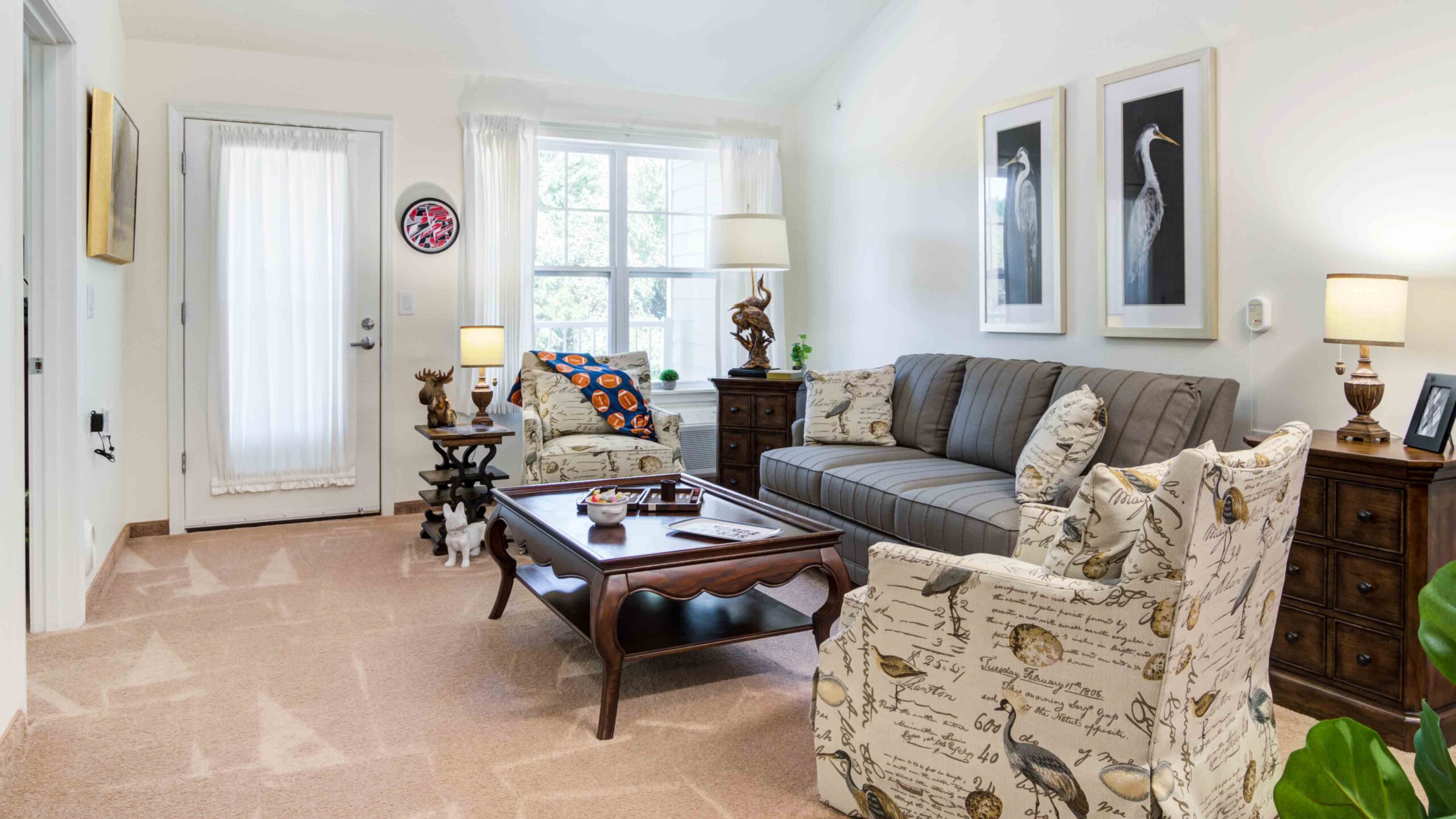 A bright living room with a gray upholstered sofa, two patterned armchairs, a wooden coffee table, side tables with lamps, bird artwork on the wall, and light streaming in from a large window and glass door.