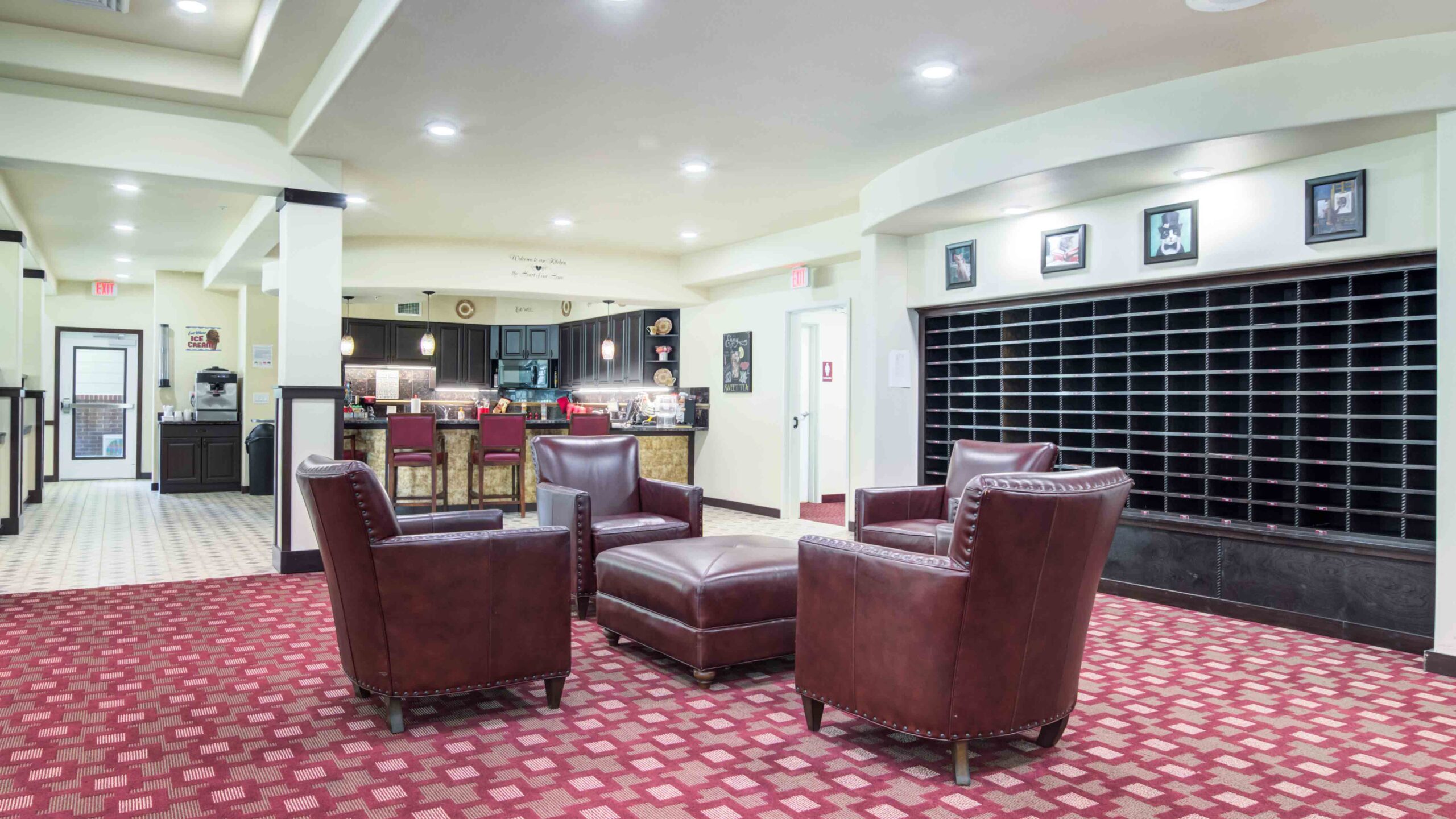 A hotel lobby with three brown leather armchairs and an ottoman on a red patterned carpet, a mail cubby system on the wall, and a reception desk with barstools in the background.