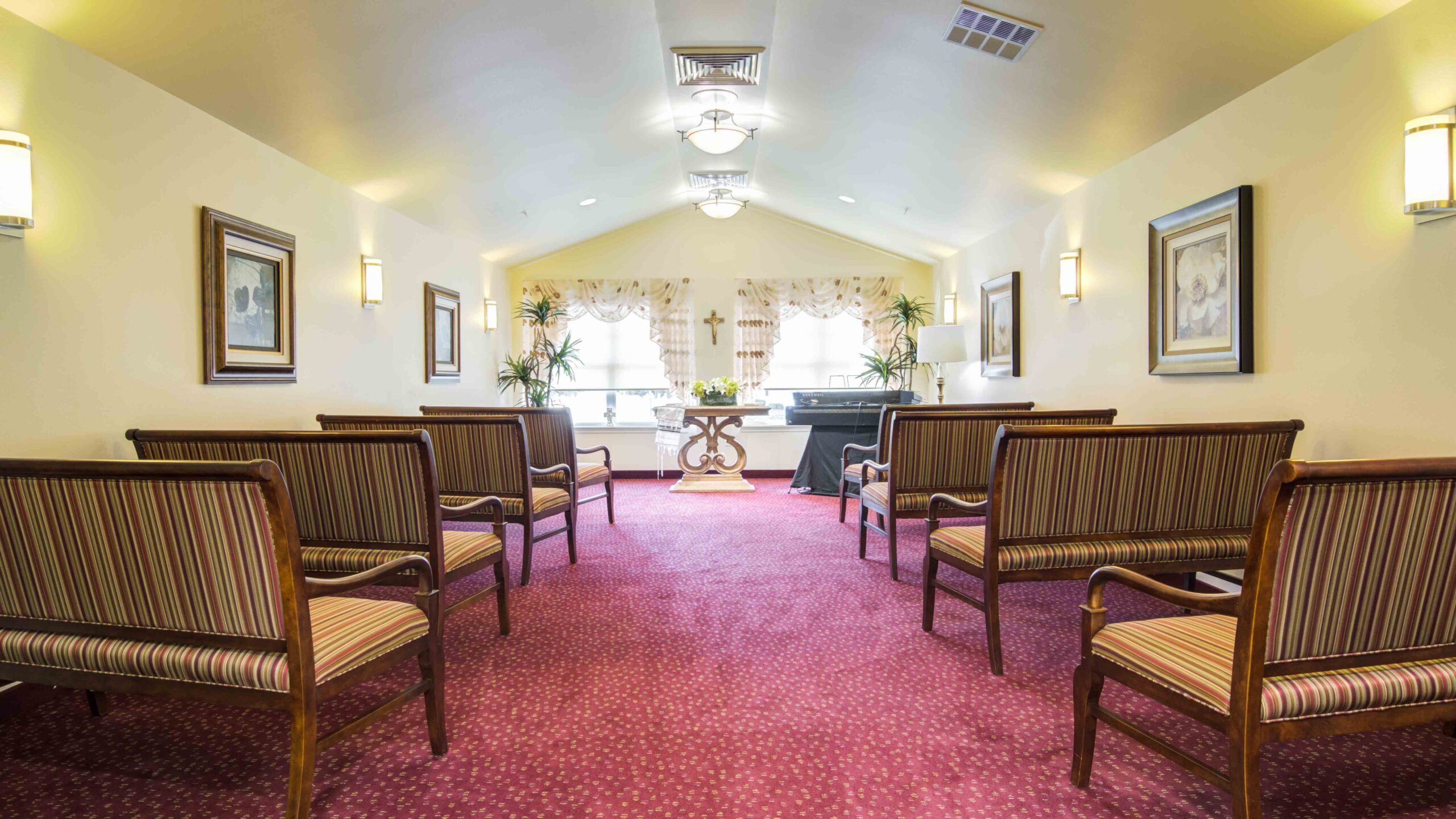 A small chapel with striped cushioned chairs facing an altar. The room has maroon carpet, framed art on cream walls, large windows with curtains, plants, and a cross on the altar. Soft overhead lighting illuminates the space.