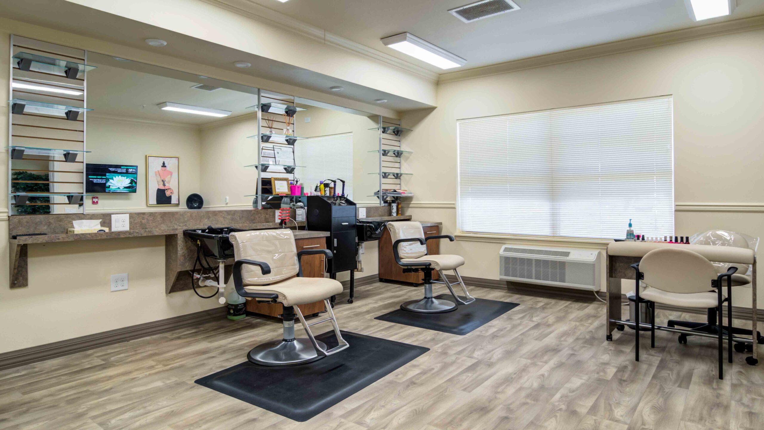 A well-lit salon with two beige styling chairs at mirrored stations, shelves holding hair products, and a small manicure table with chairs by a window. The floor has wood-like vinyl flooring.