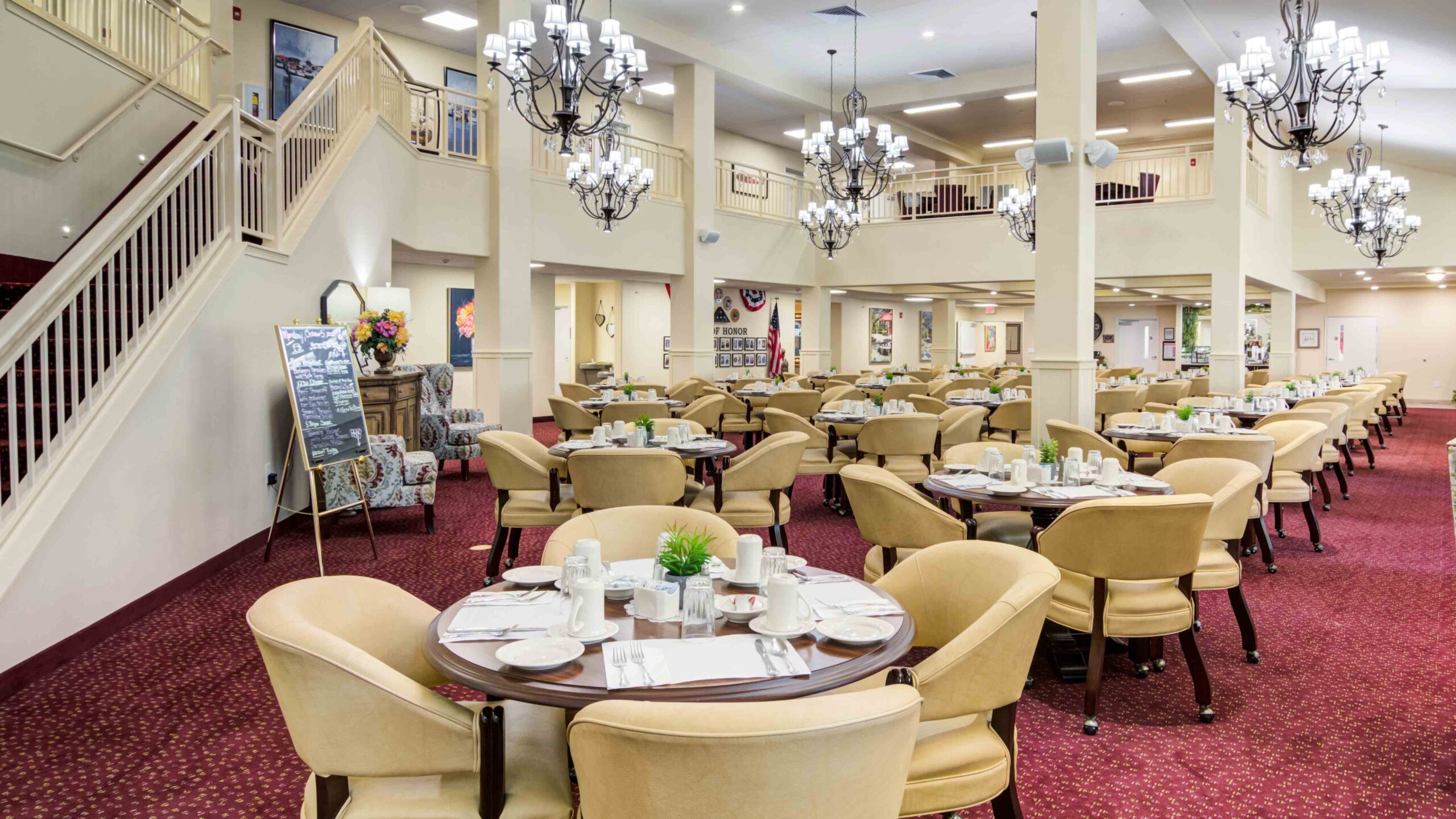 A spacious dining hall with round tables set for meals, beige cushioned chairs, red carpet, chandeliers, and a staircase leading to an upper level. Large windows and decorative accents create a welcoming atmosphere.