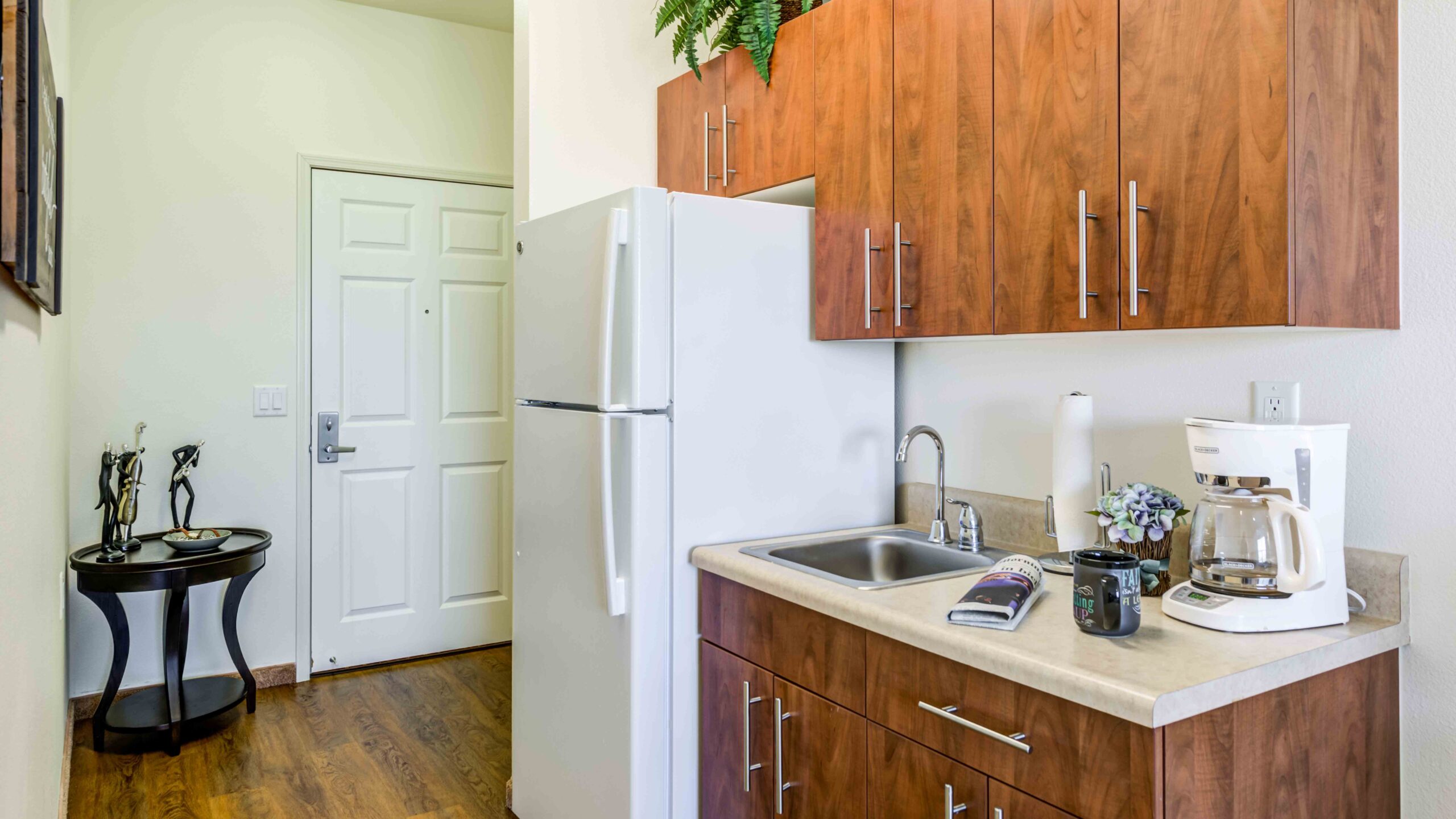 A small kitchen with wooden cabinets, a white refrigerator, and a countertop holding a coffee maker, a mug, a paper towel roll, and a magazine. The entrance door and a small side table are visible in the background.