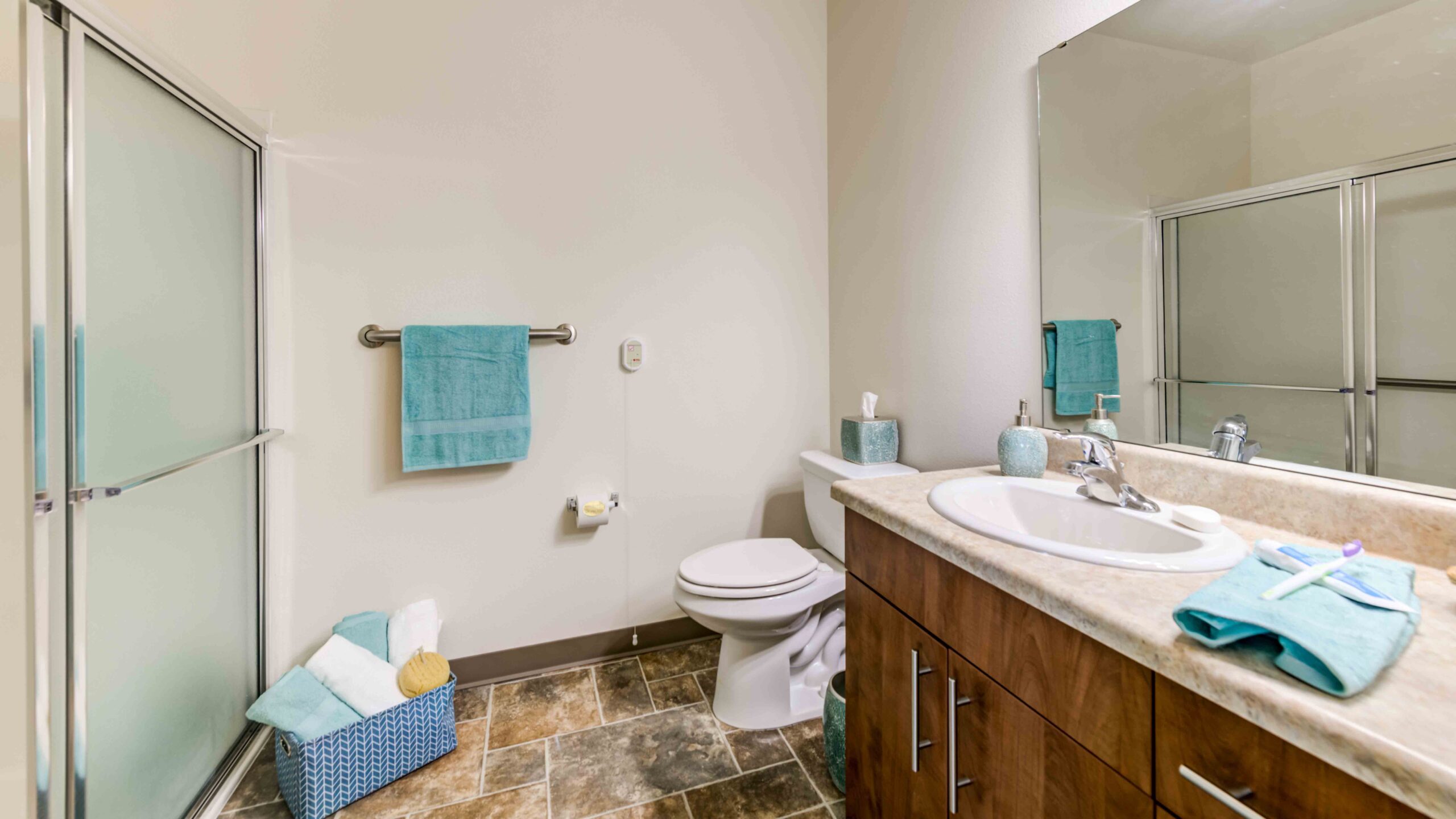 A clean bathroom with a toilet, a wooden vanity with a sink, a large mirror, a glass shower, and teal towels and accessories. A blue basket with towels sits on the floor near the toilet.
