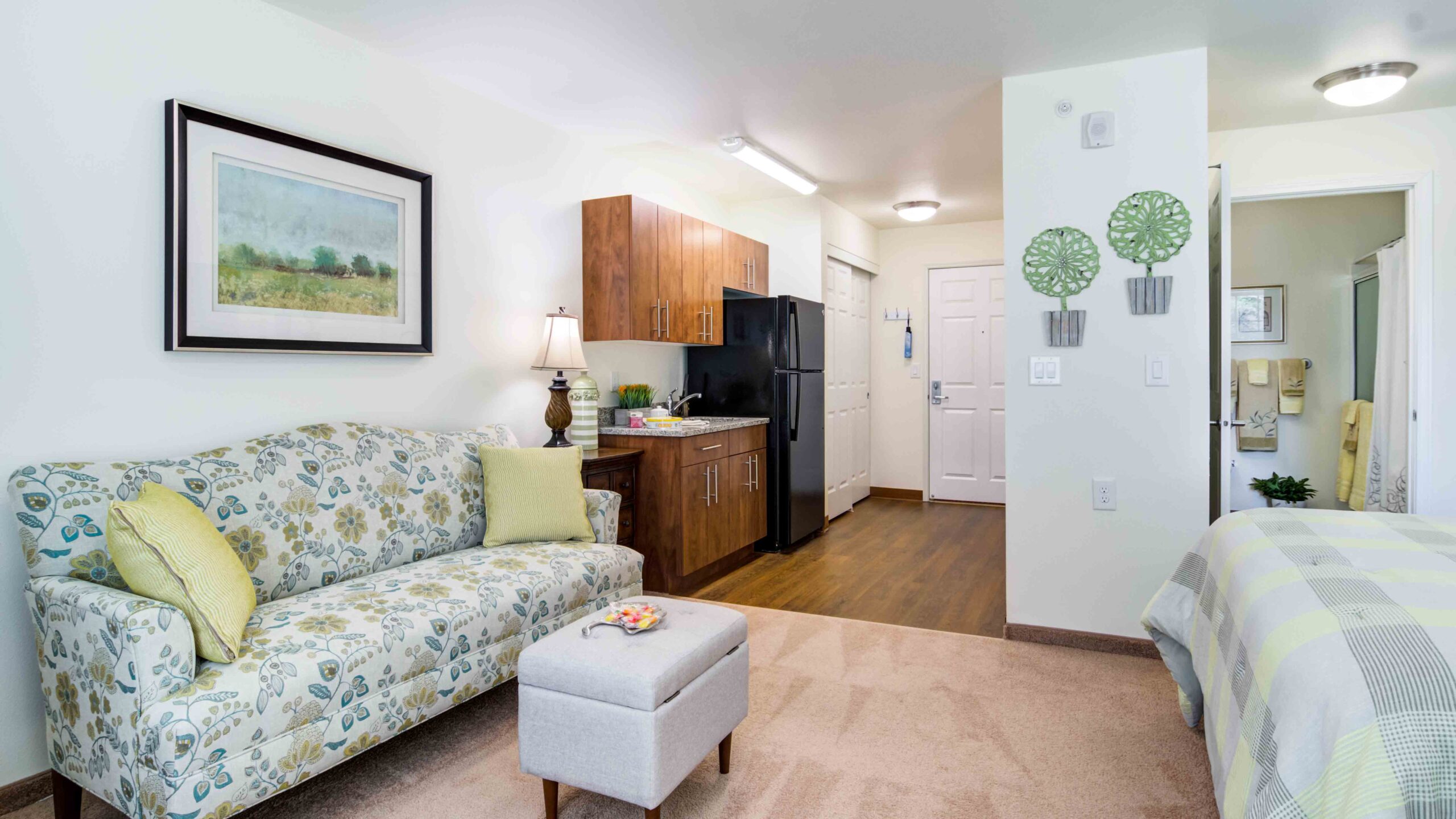 A bright studio apartment with a floral sofa, ottoman, and lamp, adjacent to a compact kitchen with wooden cabinets, a black fridge, and an entry door; a bed and bathroom are visible in the background.