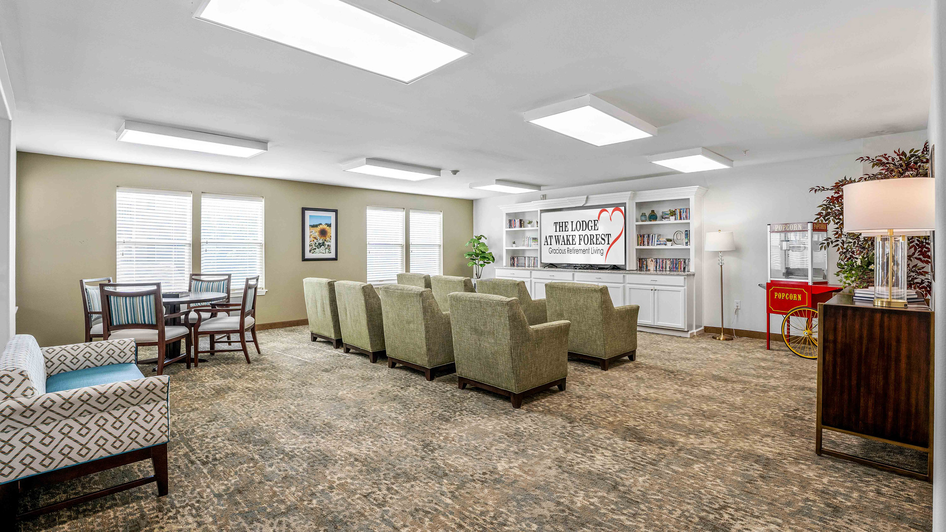 A bright lounge with several green armchairs facing a TV, bookshelves, and a popcorn machine. There are large windows, framed art, a dining table with chairs, and potted plants. The room has beige carpet and overhead lights.
