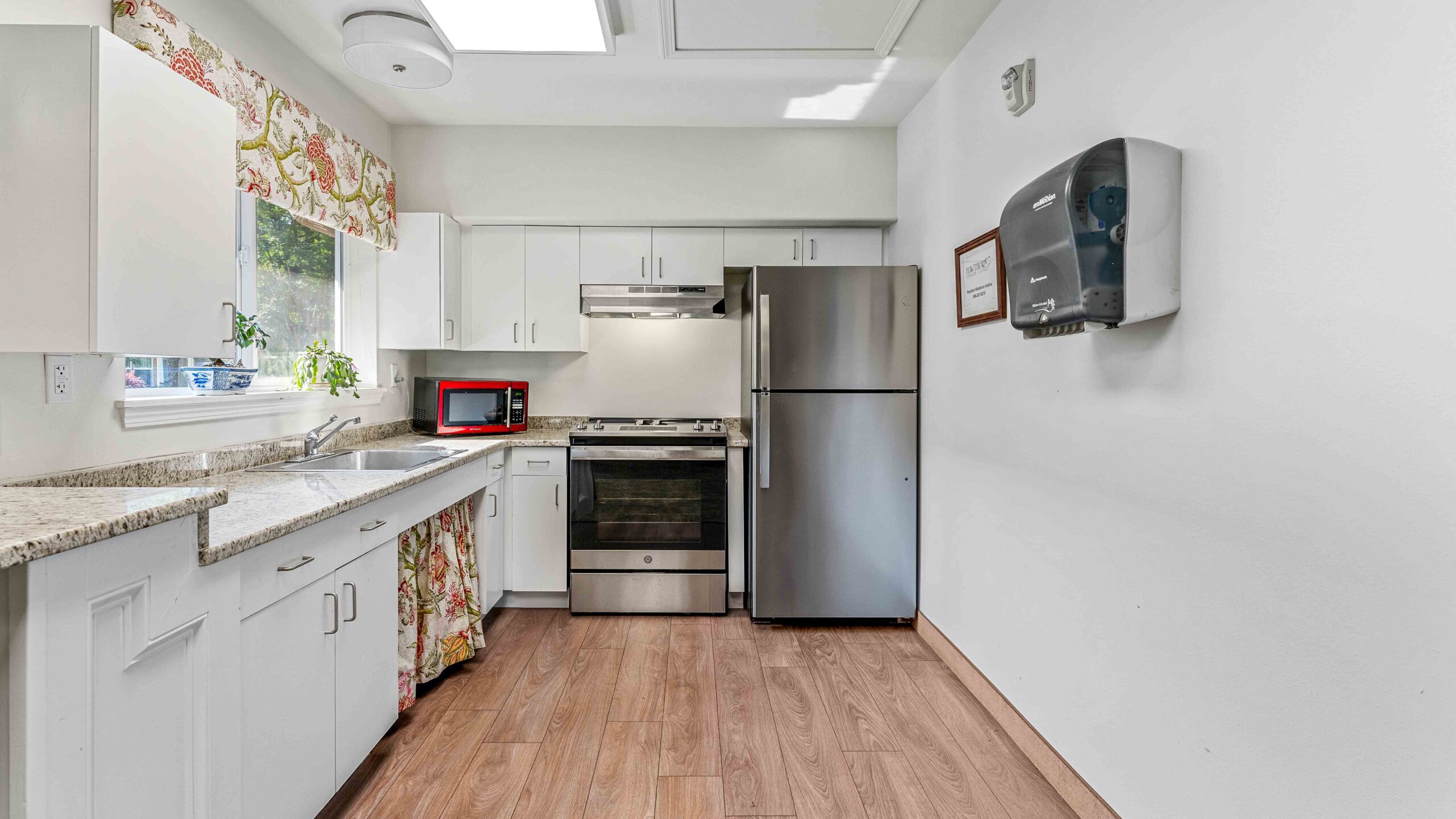 Modern kitchen with white cabinets, stainless steel appliances including a fridge and stove, a red microwave on the counter, granite countertops, floral curtains, and wood-style flooring.