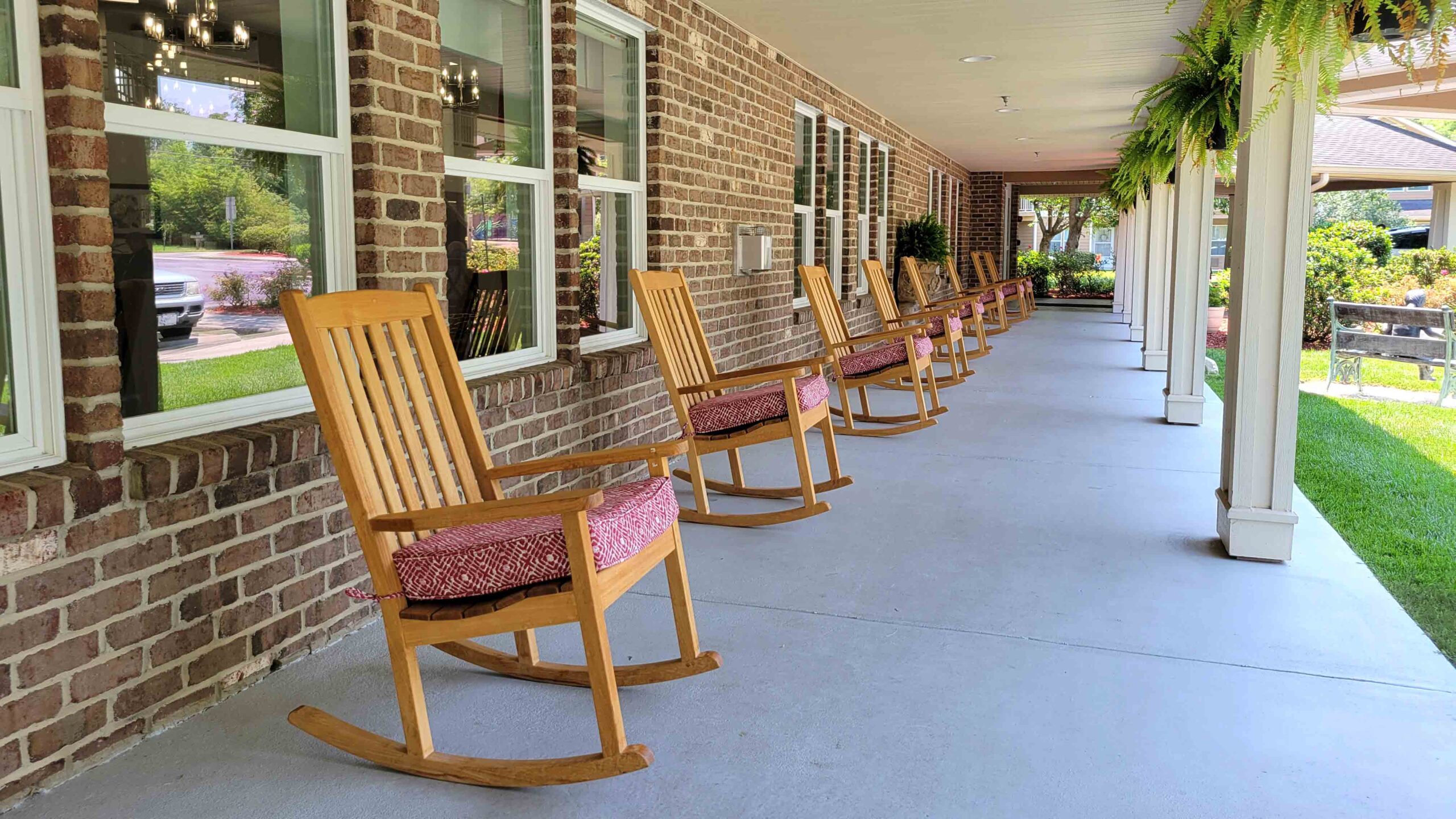 A row of wooden rocking chairs with red cushions lines the covered porch of a brick building, overlooking a green lawn and hanging ferns.