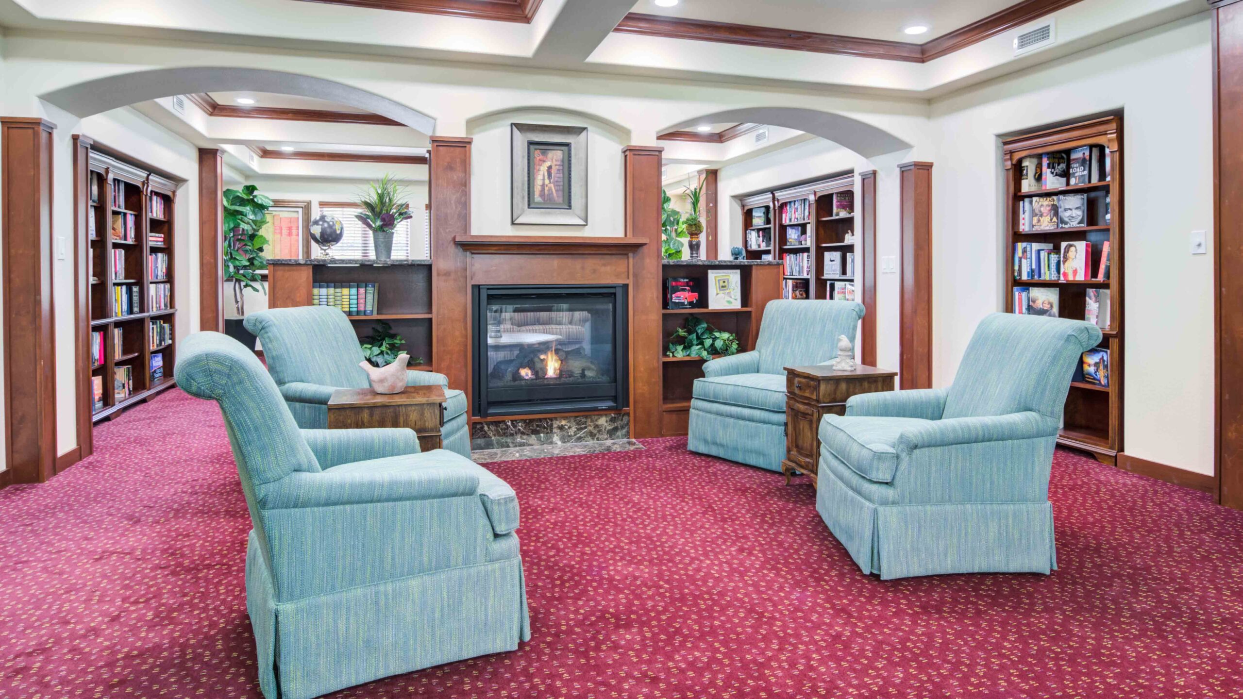 A cozy library lounge with four blue armchairs arranged around a fireplace. Bookshelves line the walls, and the room has red carpet, wooden trim, green plants, and framed art above the mantel.